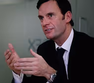 Man in a black suit and white shirt gesturing with his hands while speaking.