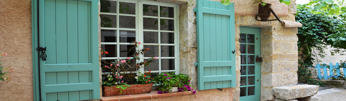 A photo of a house in France with flowers in the window ledges. The doors and windows of the property are painted in a teal blue. 